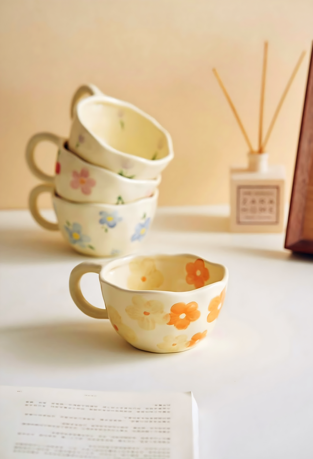 a cream based mug with yellow and orange flower pattern in the foreground next to a book, in the background three similar mugs but with different patterns of red cherry, blue flowers, pink flowers and purple tulip stacked, next to a air diffuser