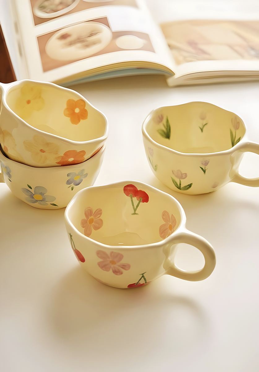 a ceramic cream based mug with red cherry and pink flowers pattern on a surface with three other similar mugs but in different pattern like blue flowers, yellow and orange flowers and purple tulips behind, in the background, a cooking book laying around