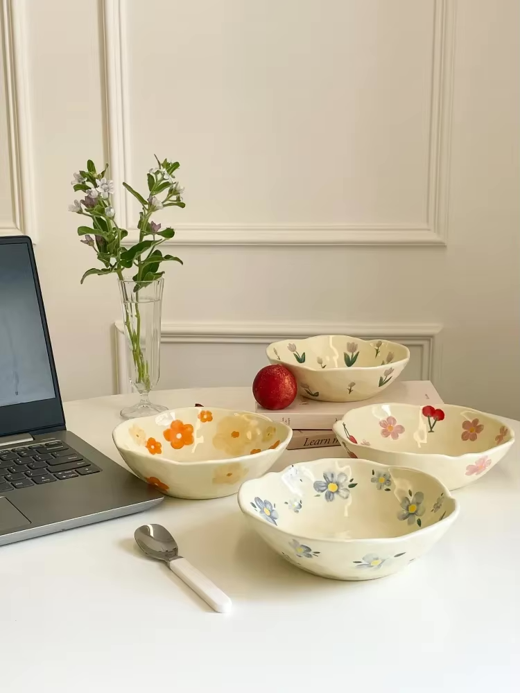 a red cherry pink flower patterened, a purple tulip patterned, a orange flower patterned and a blow flower patterned bowls on a white table, next to them is a glass vase of flowers and a laptop