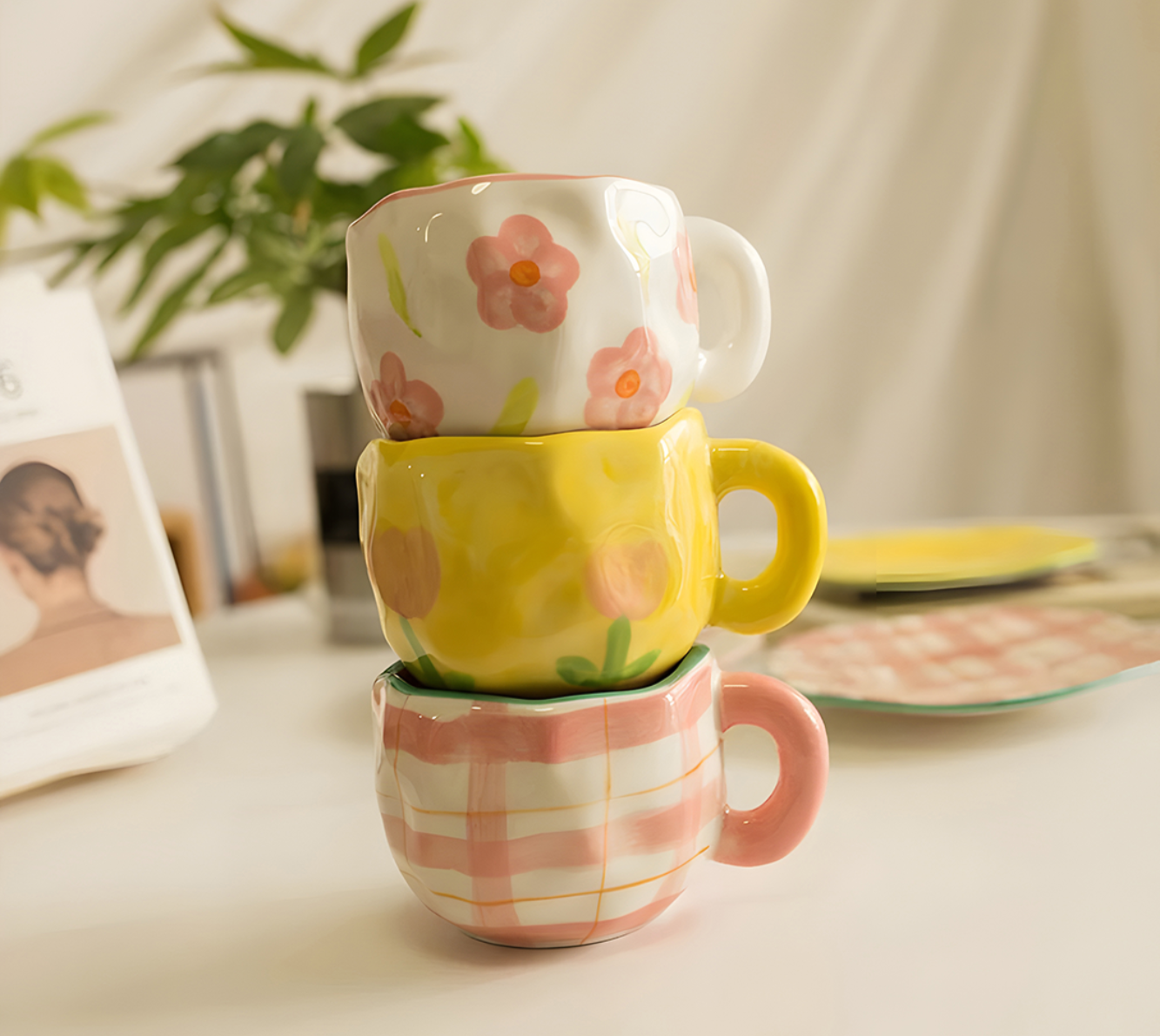 ceramic pink plaid mug stacking with pink flower mug and yellow tulip mug on a white table with green plants in the background, aesthetic display