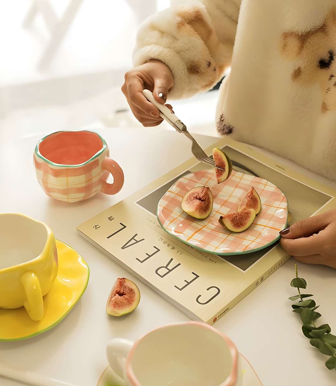 a person using pink plaid ceramic saucer eating figs. Pink plaid mug is next to her hand, with a book underneath on a white table.