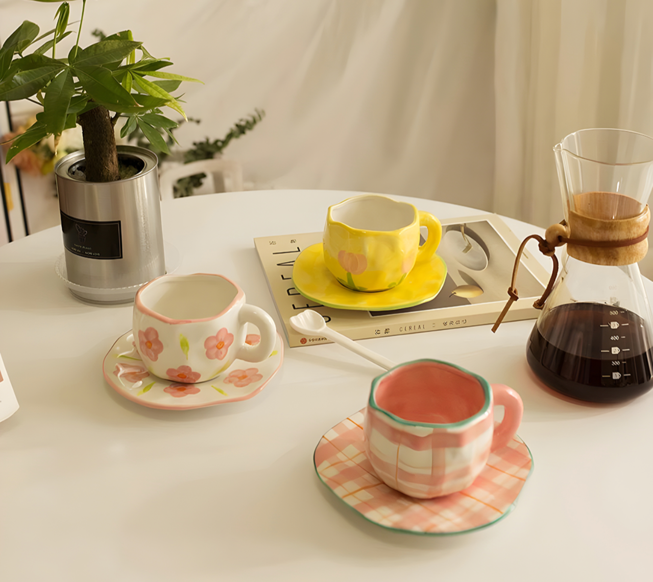ceramic pink plaid mug with pink flower mug and yellow tulip mug on a table aesthetic display, coffee pot on the side with green plants.