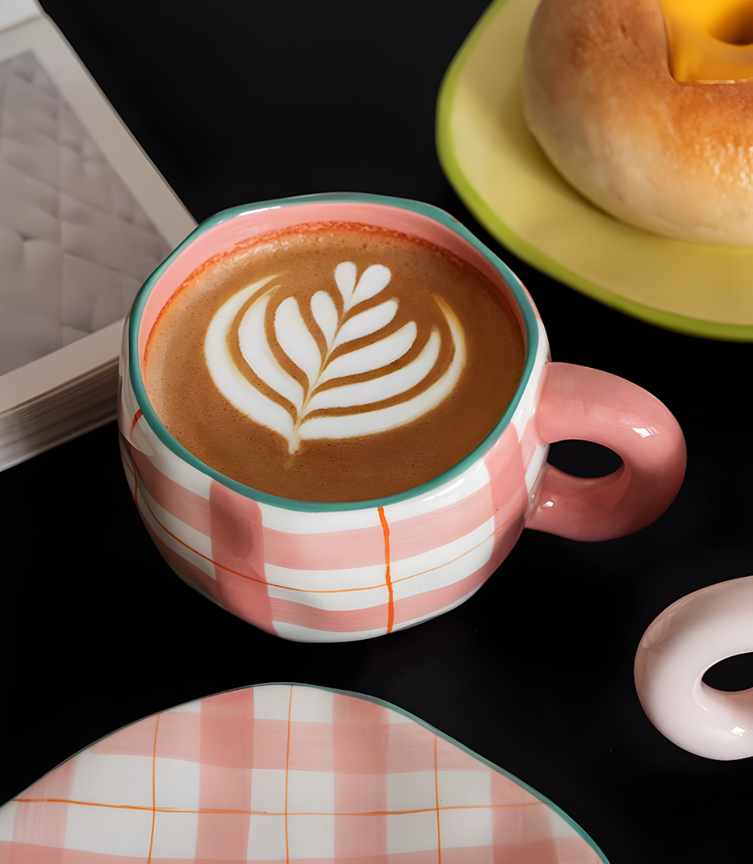 latte art in a ceramic pink plaid mug with a bagel next to the mug