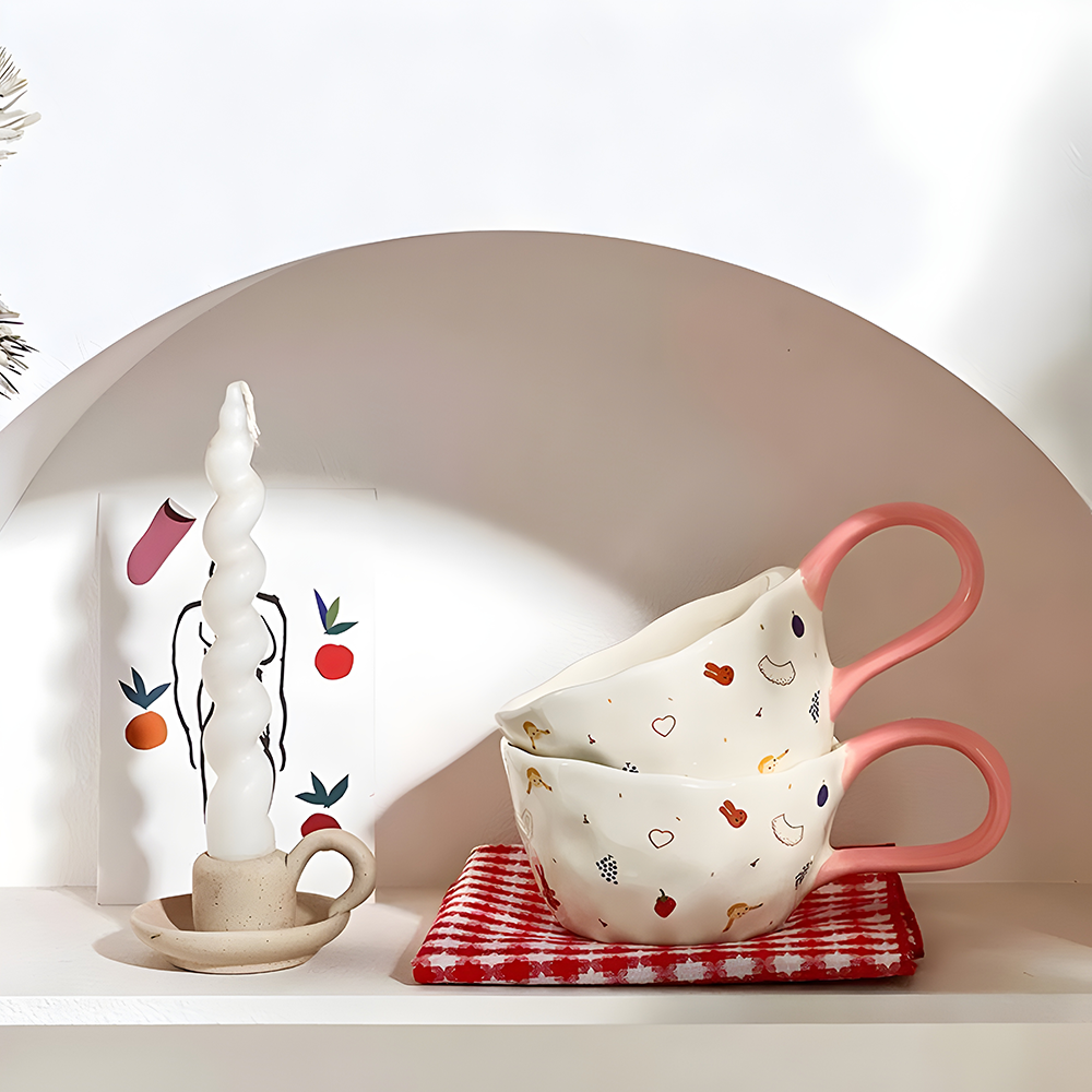 2 cream based ceramic mugs with little girl, red cherry, heart, strawberry, bunny and grape print, a big loop pink handle illustration, stacking on top of a red and white plaid tea towl, placed inside a shelve with a white candle next to them