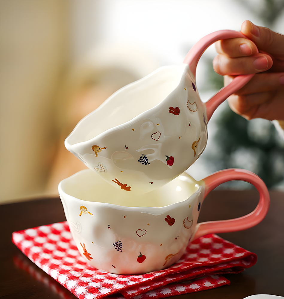 a hand holding a cream based ceramic mug with little girl, red cherry, heart, strawberry, bunny and grape print, a big loop pink handle in the air; underneath is a same mug on top of a red and white plaid tea towel, all on top of a wooden table