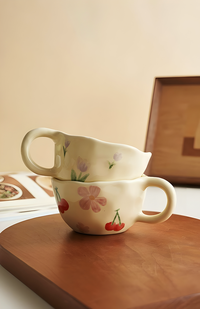 a ceramic cream based mug with red cherry and pink flowers pattern stacking with a similar ceramic mug but in purple tulip pattern, on a wood table with cooking books in the background