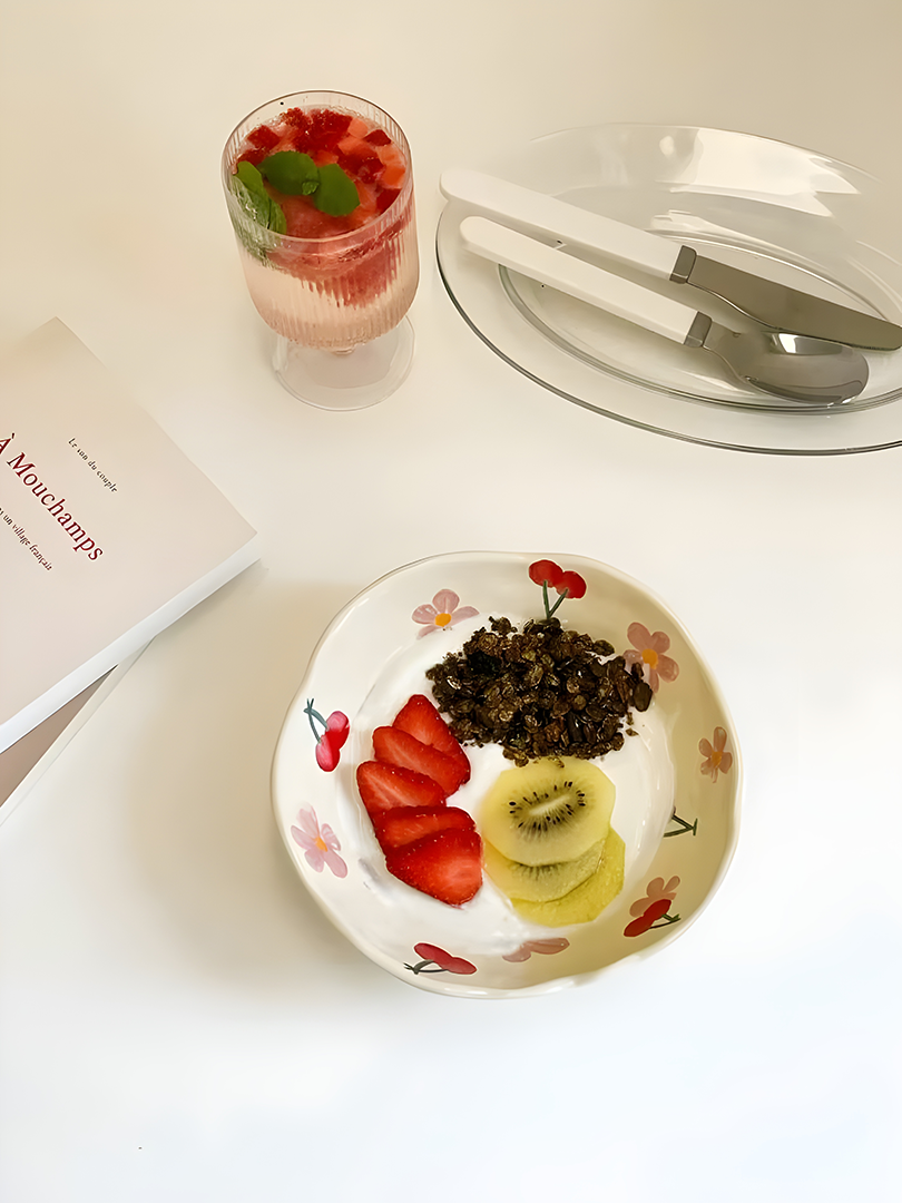 red cherry pink flower cream based ceramic bowl filled with yogurt, strawberries, kiwi, oats; a glass of sparkling drink with strawberries and mint next to it; A glass dish with fork and spoon and a book surronding the ceramic bowl