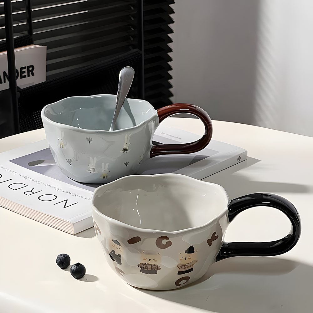 a grey bear and letter patterned ceramic mug with black big loop handle in the foreground, a blue bunny and tulip patterned ceramic mug with brown big loop handle in the background, on top of a magazine, on a white table