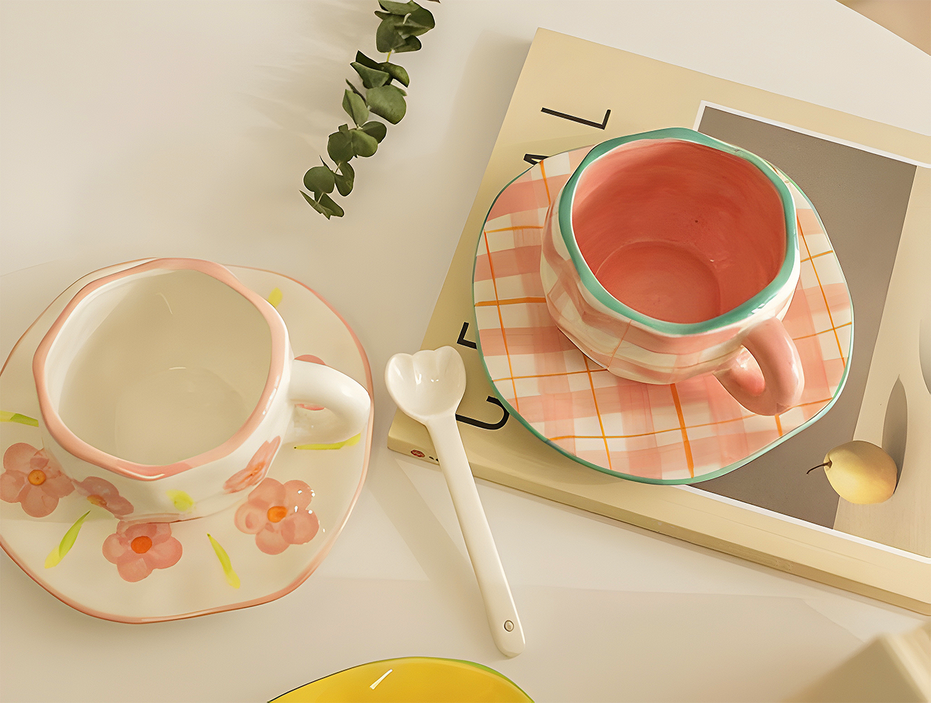 a pink plaid ceramic mug with a saucer on top of a book, a spoon next to it and  a pink flower mug with saucer