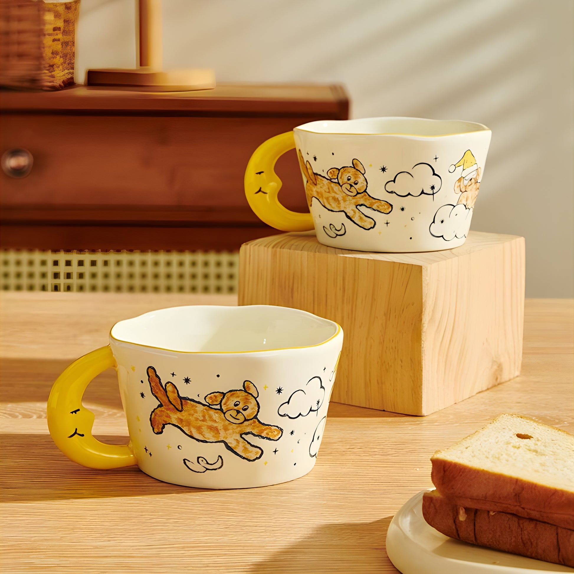 2 moon shaped handle cearmic mugs with blue moon, cloud, a dog and stars placed on a wooden surface with warm light shining. A toast in a dish in the foregrond. 